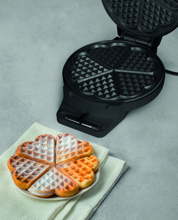 Homemade heart-shaped Belgian waffles dusted with powdered sugar, served next to an open electric waffle iron against a light gray background.