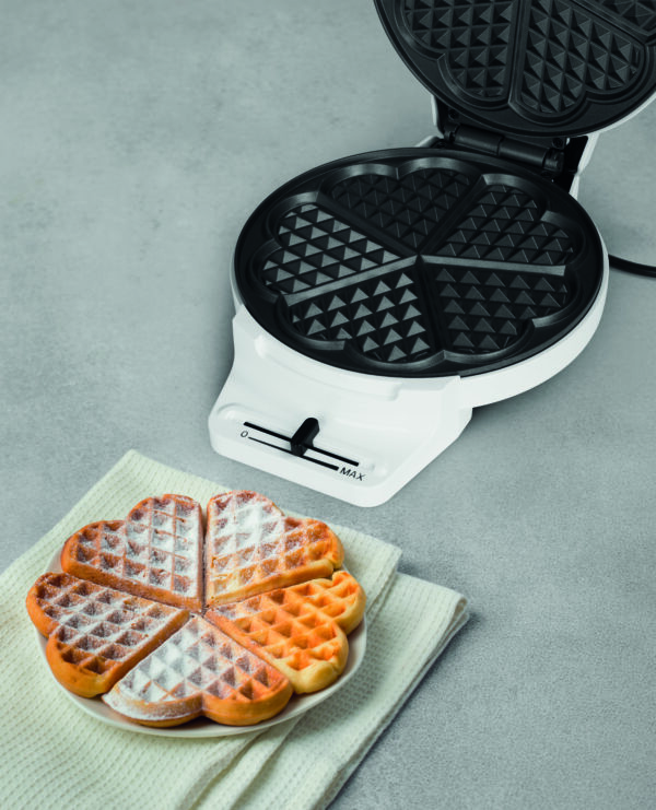 Homemade heart-shaped Belgian waffles dusted with powdered sugar, served next to an open electric waffle iron against a light gray background.